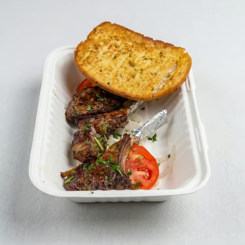 Grilled lamb chops with garlic bread, tomatoes, and onions in a white takeout container.