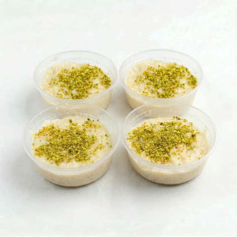Four clear plastic cups of rice pudding, topped with finely chopped green pistachios, on a white surface.